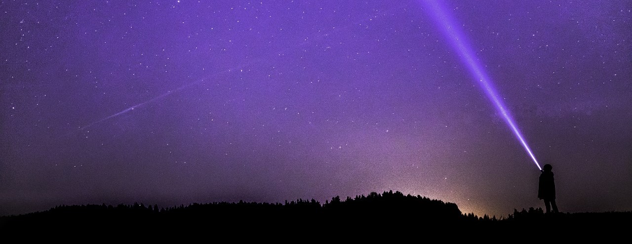 Vast night sky with bright stars, and a tiny person in silhouette holding up a flashlight
