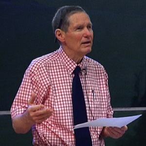 Older man in a checked shirt, holding a page and presenting to an audience
