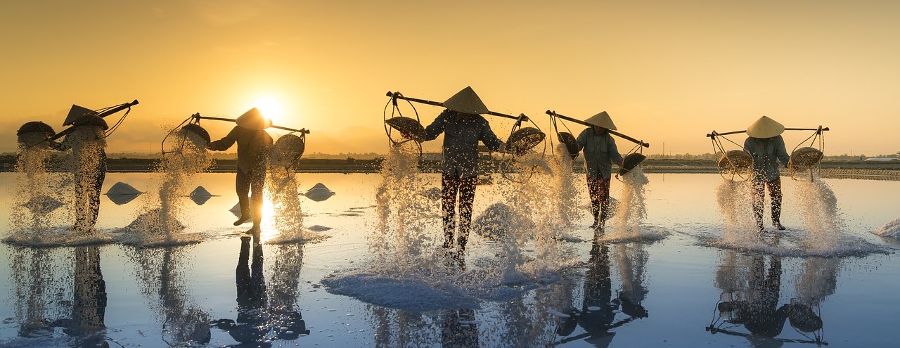 Four adults shown in silhouette, with pails balanced across both shoulders, walking through shallow water