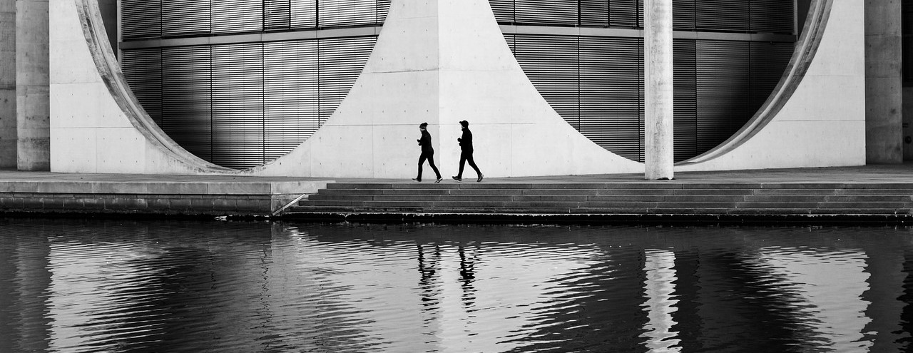Black and white photo of a modern architectural structure overlooking a body of water edged by stone steps. Two people walk along the bank.