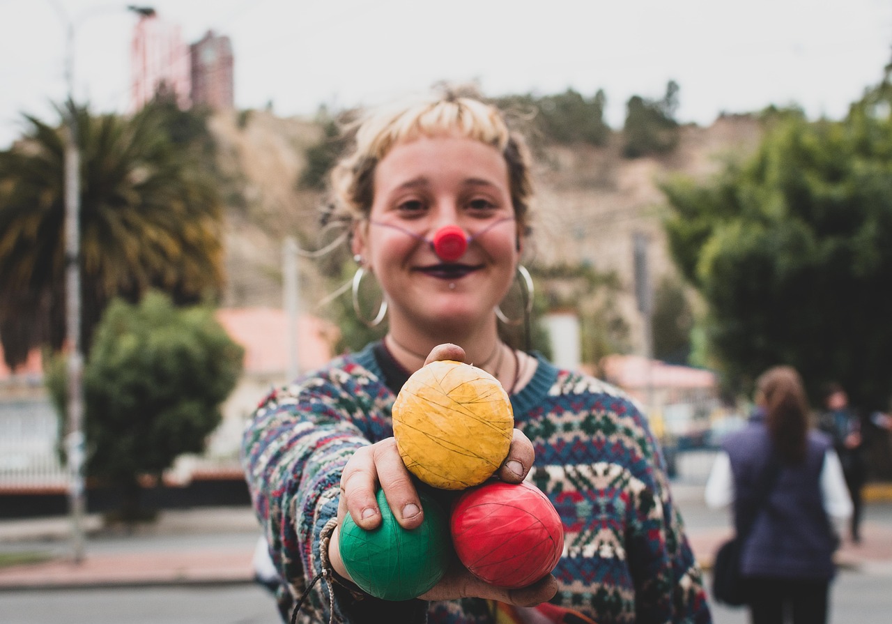 A woman in colourful clothing, wearing a red nose, holding out three coloured juggling balls