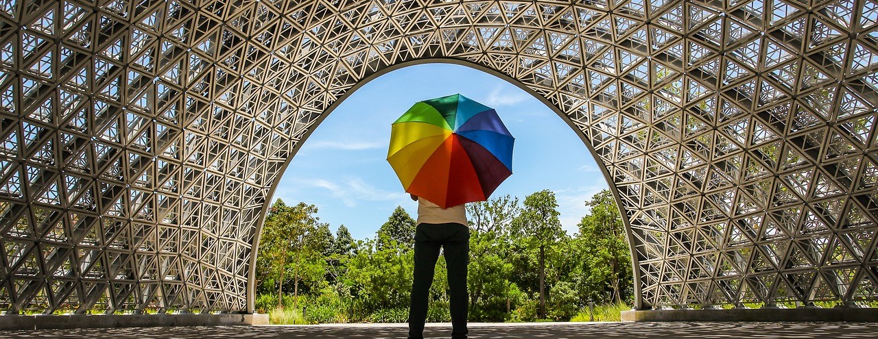 A person standing under a walkway, holding up a rainbow umbrella.